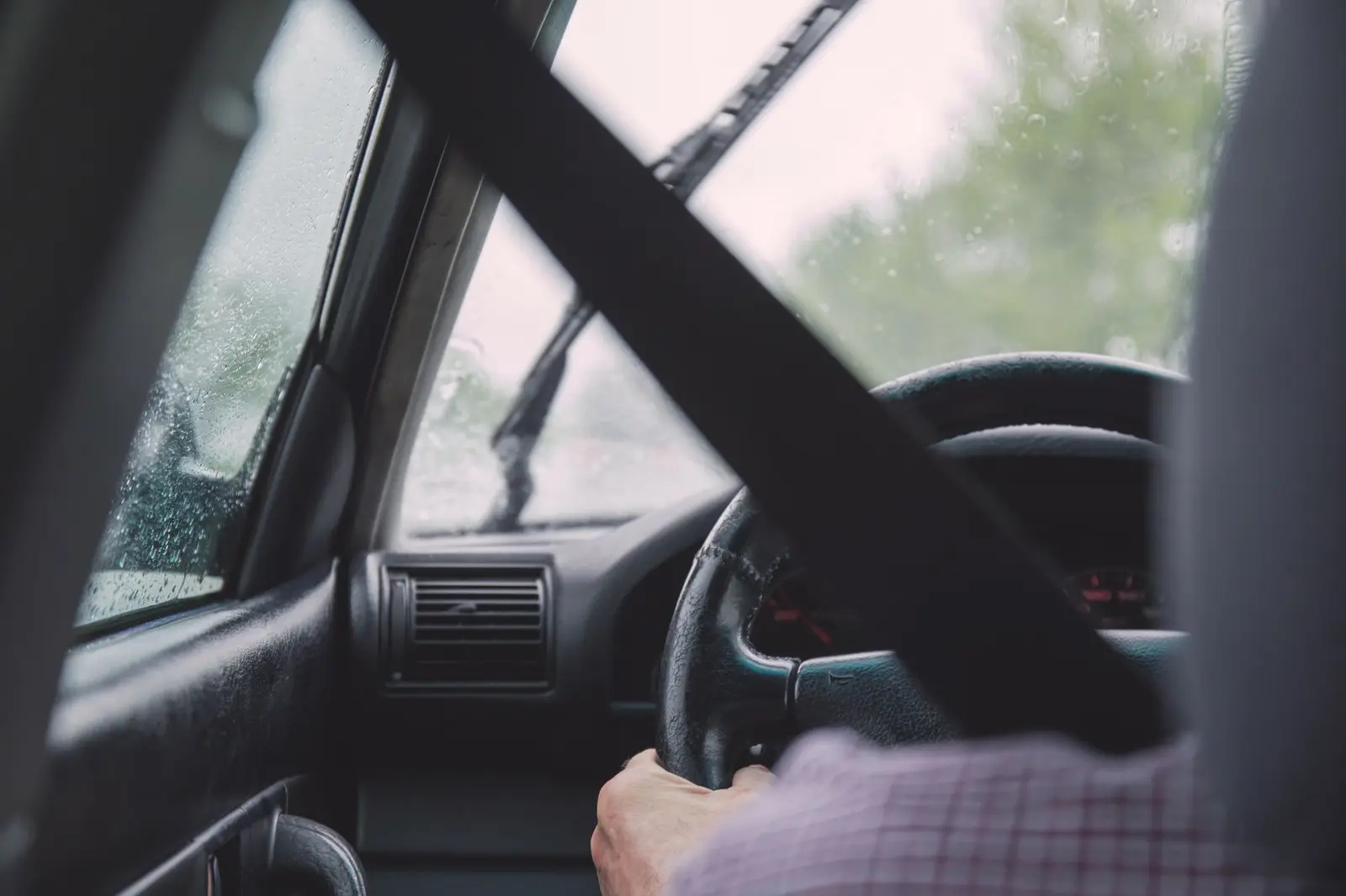 A driver holding the wheel, with a seat belt across the chest, in rain