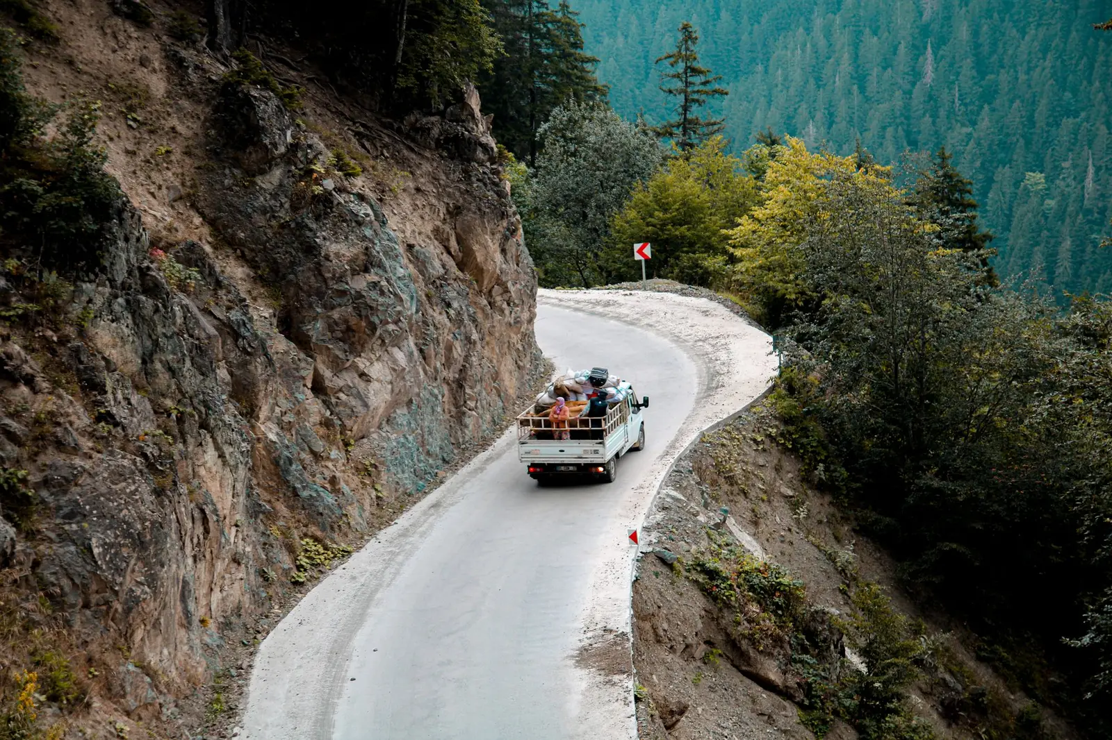 A small goods vehicle on a narrow mountain road cut into a forested slope